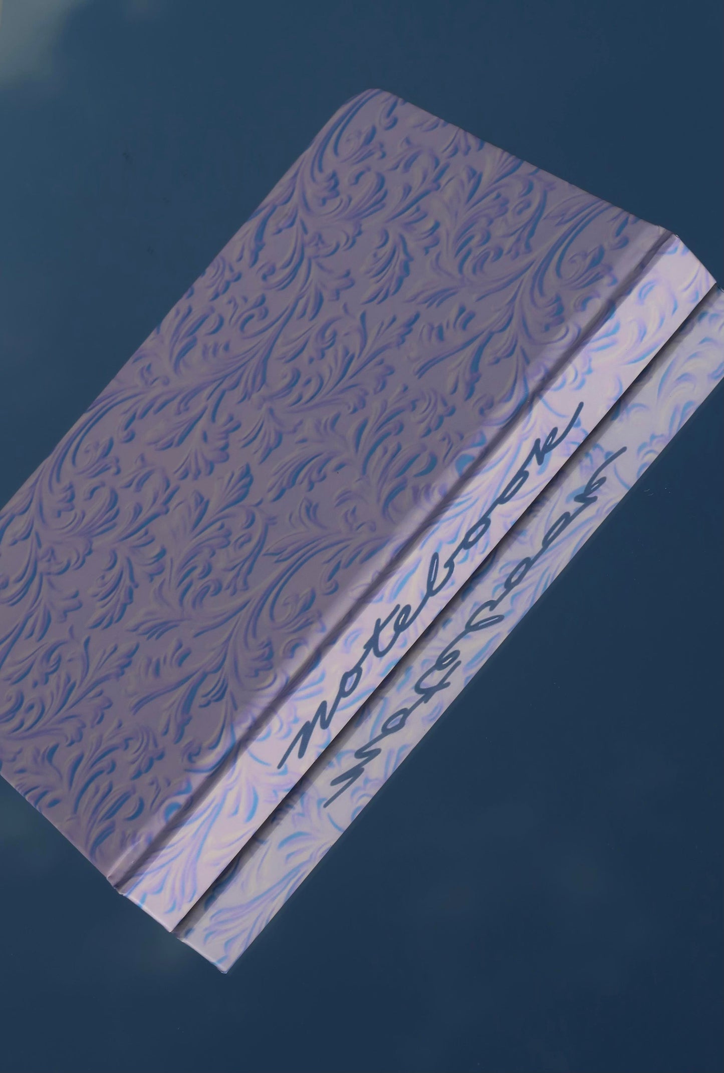 blue and purple hardcover journal with a beautiful designed pattern appears to be floating in the sky and has a reflection with the spine of the notebook spelling out "notebook" in cursive
